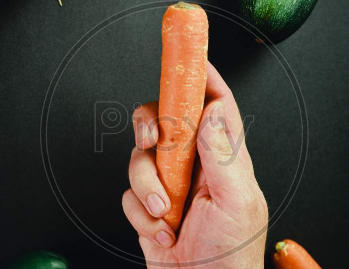 Hand Grabbing A Carrot Surrounded By Vegetables With A Black Table