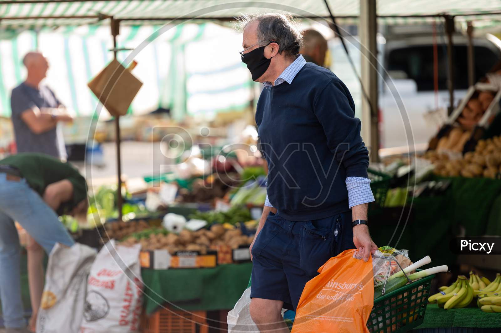 A Mature Man Wearing A Protective Face Mask Carries Shopping At An Outdoor Market