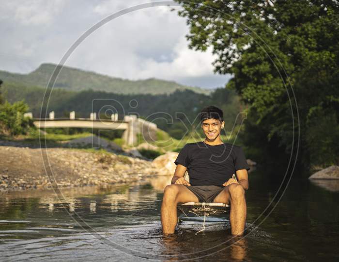 A Young Handsome Asian Boy Sitting In A Chair In The Middle Of A River, Golden Hour.