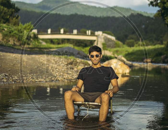 A Young Handsome Asian Boy Sitting In A Chair In The Middle Of A River With Black Sunglasses, Golden Hour.
