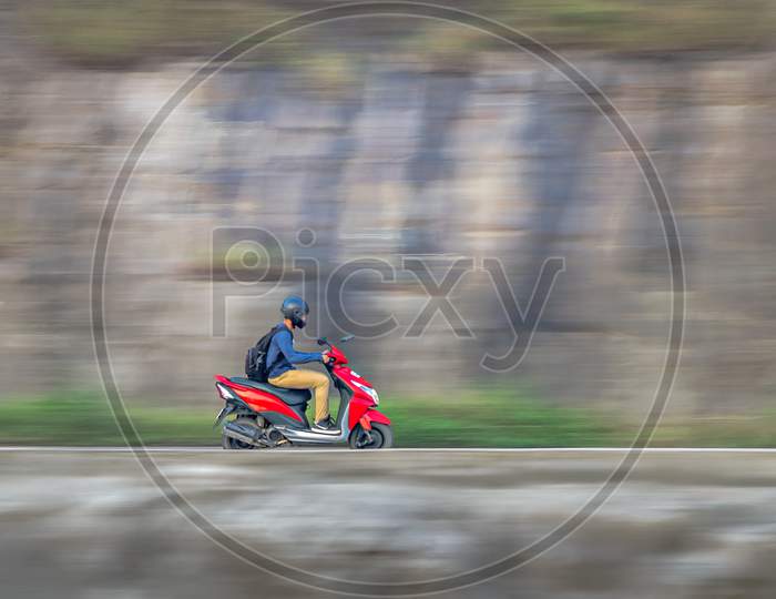 Motion Blur Image Of A Rider Wearing Helmet For Safety, Riding Uphill On A Red Two Wheeler Moped.