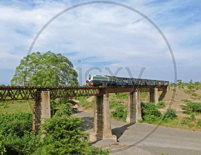 A Small Narrow Gauge Train With Diesel Locomotive Crossing An Old Stone And Iron Bridge In Chamak, Maharashtra, India.