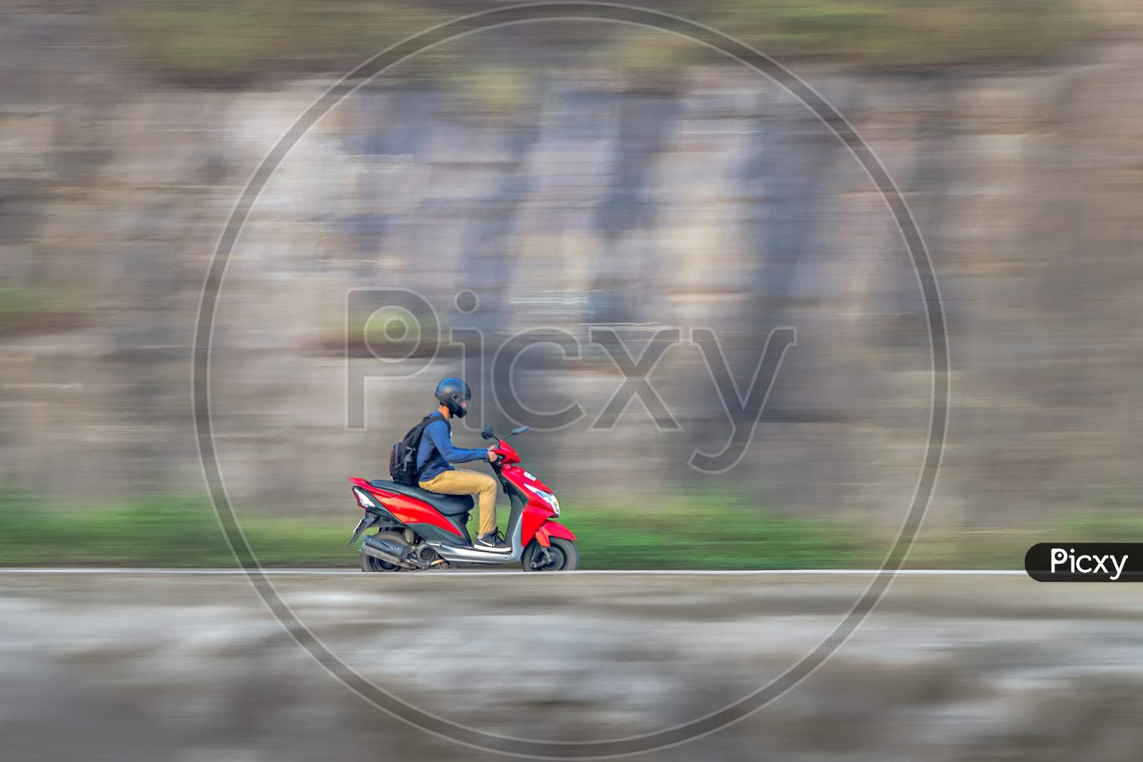 Motion Blur Image Of A Rider Wearing Helmet For Safety, Riding Uphill On A Red Two Wheeler Moped.