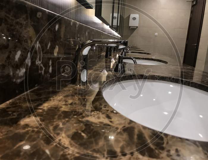 An Luxury Wash Room Or Rest Room Interiors Such As Beveled Type Mirror And Polished Marble Counter Top Wash Basin And Faucet Setup For An Male Toilet Area Of An Shopping Mall Or An Five Star Hotel