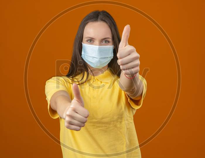 Young Woman In Yellow Polo Shirt And Medical Protective Mask Showing Thumb Up Sign Looking At Camera With Positive Expression On Orange Background