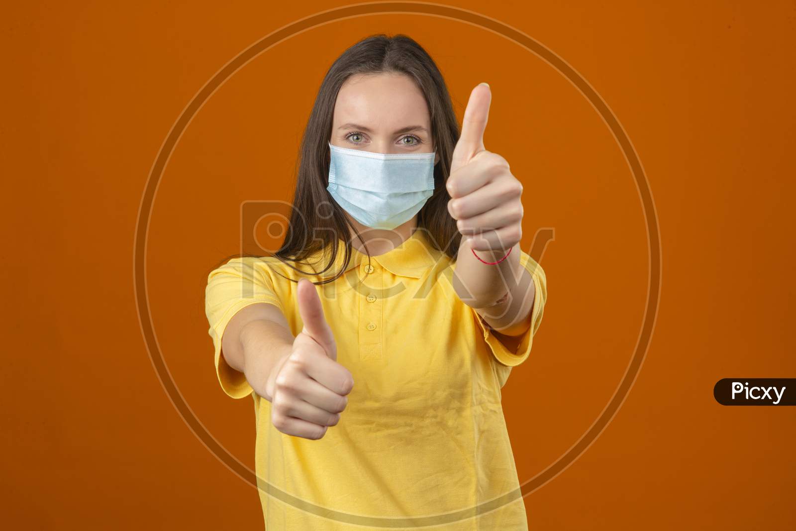 Young Woman In Yellow Polo Shirt And Medical Protective Mask Showing Thumb Up Sign Looking At Camera With Positive Expression On Orange Background