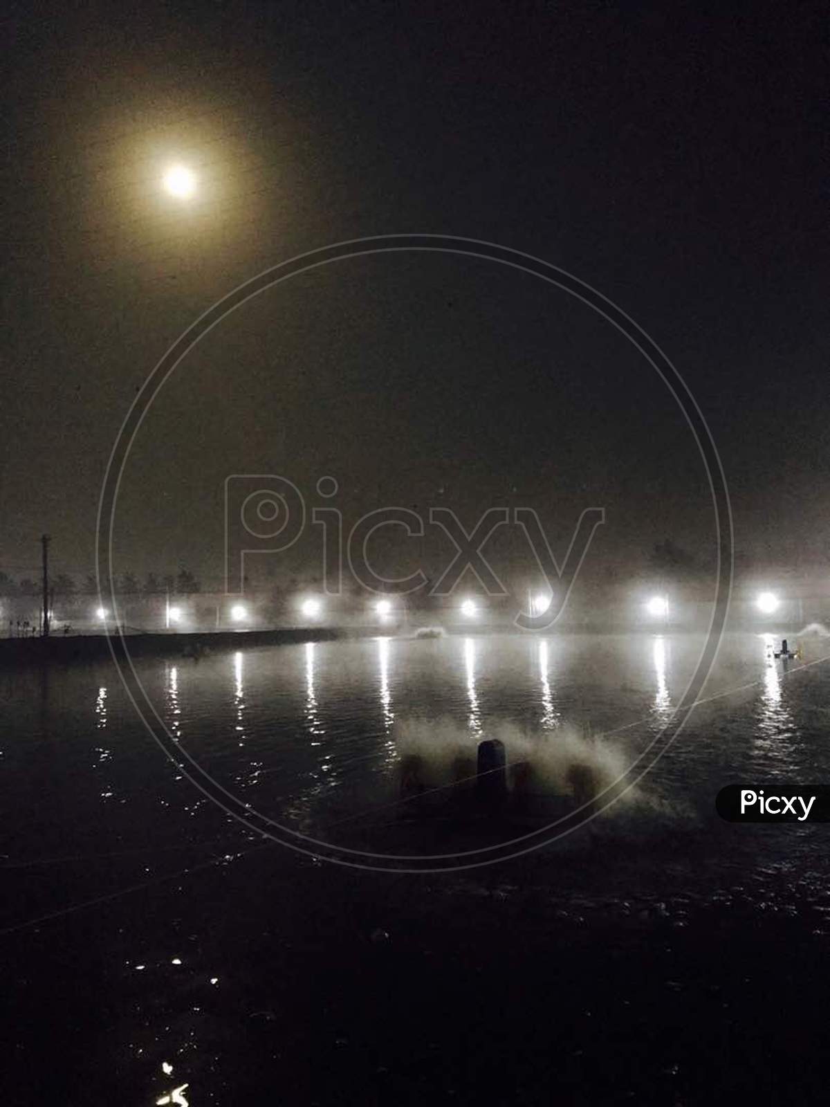 A row of lights with Moon reflecting in aquaculture pond water and with areators at night time