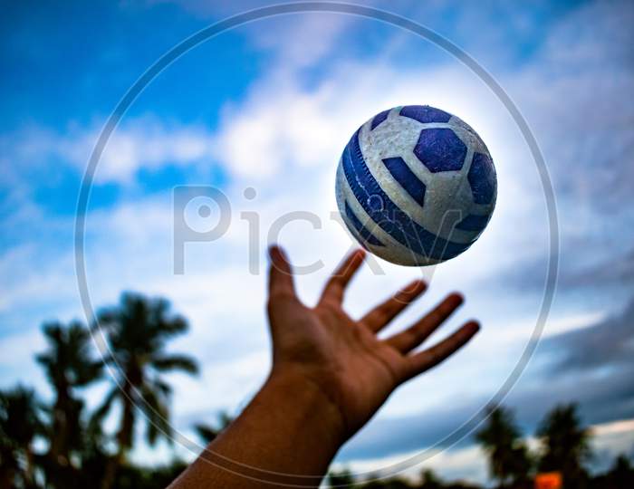 A Man Throwing Ball By Hand Against The Clear Blue Sky With White Clouds. Close up macro shot of plastic cricket ball float in air on blur nature background. Creative Photography, Copy Space For Text.