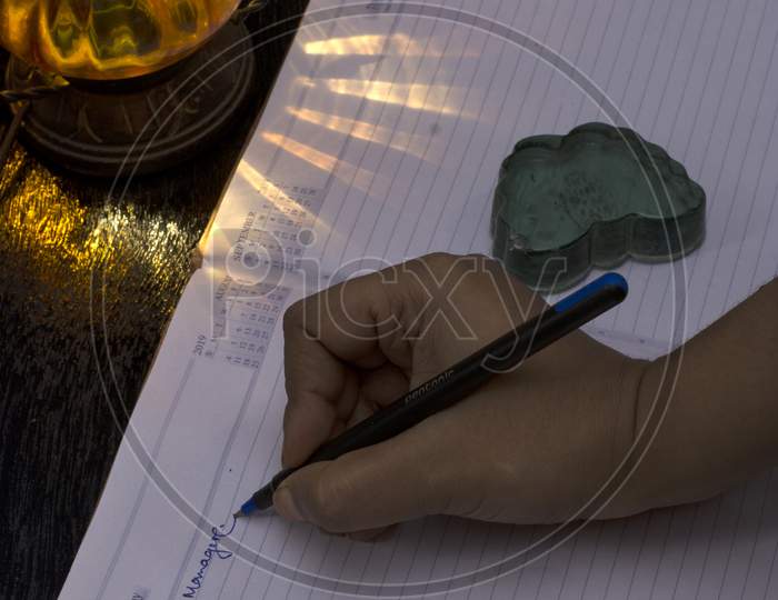 A Hand Writing Something On An Office Notebook With Pen Eye Glasses With A Lamp Shade