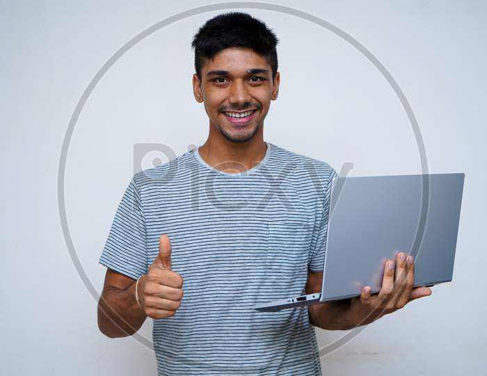 `Young Indian Handsome Boy Showing Thumbs Up And Smiling While Looking Into The Camera, Holding Laptop In His Other Hand.
