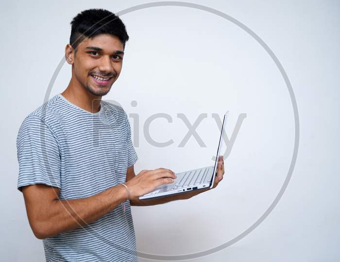 Young Indian Handsome Boy Smiling Into The Camera While Holding His Laptop In His Arms.