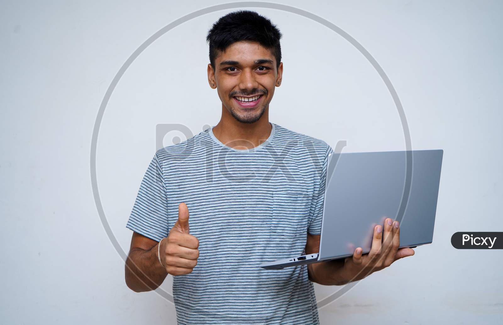 `Young Indian Handsome Boy Showing Thumbs Up And Smiling While Looking Into The Camera, Holding Laptop In His Other Hand.