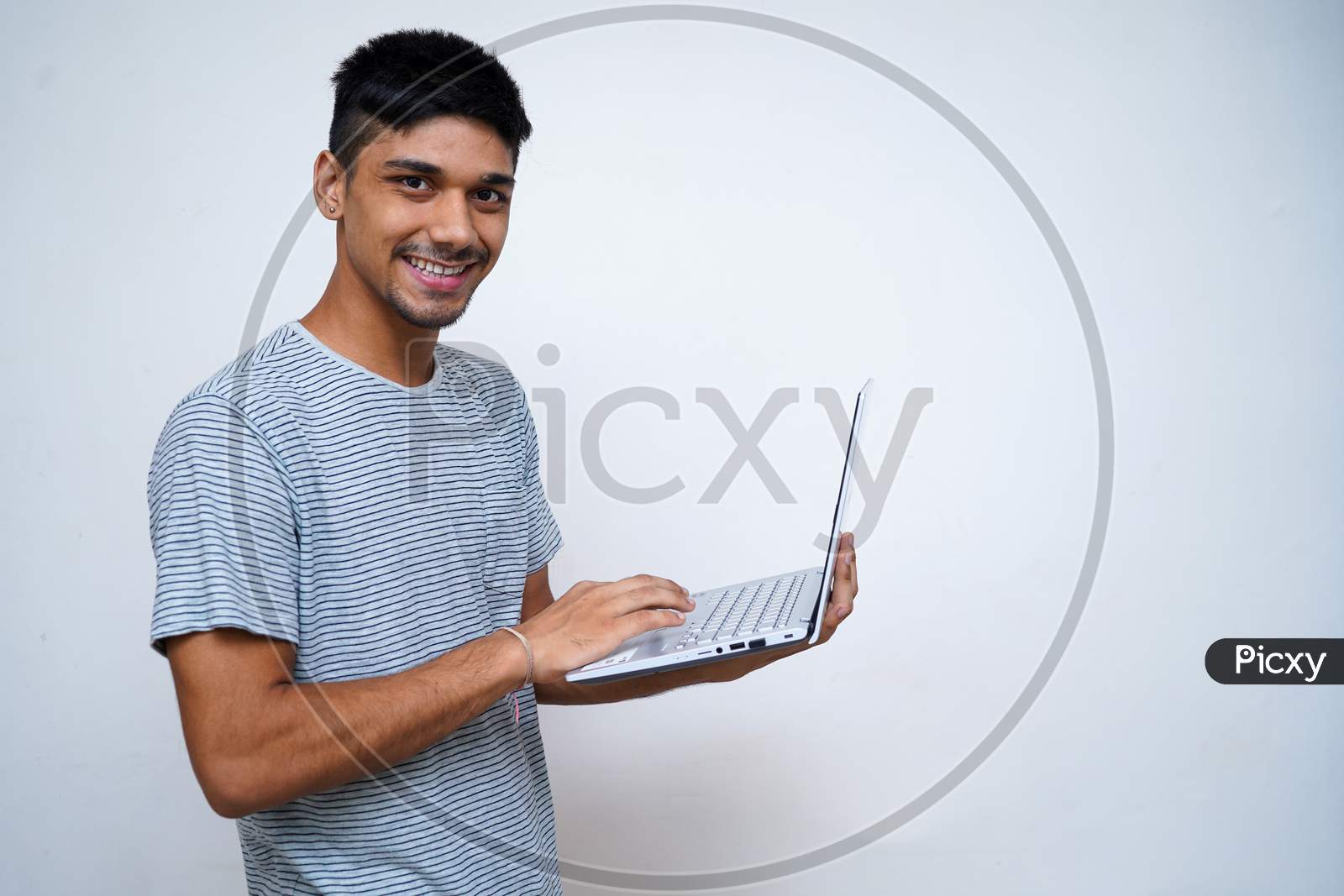 Young Indian Handsome Boy Smiling Into The Camera While Holding His Laptop In His Arms.