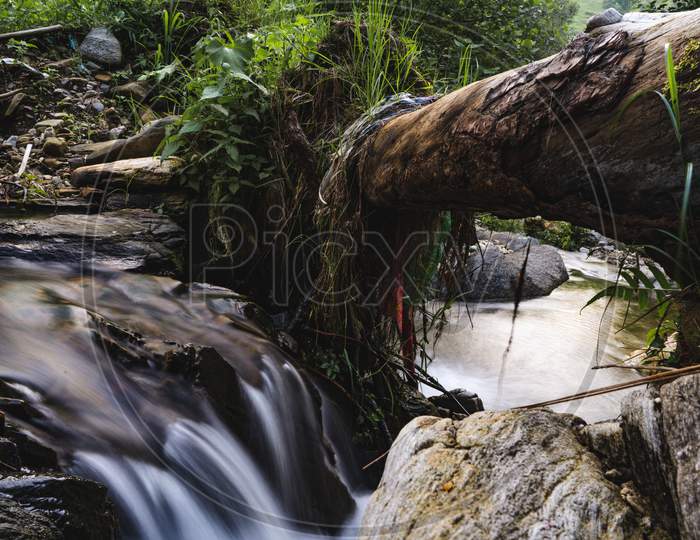 Slow Shutter Photo Of A Flowing River With A Broken Log On The Top Of It Forming A Bridge.