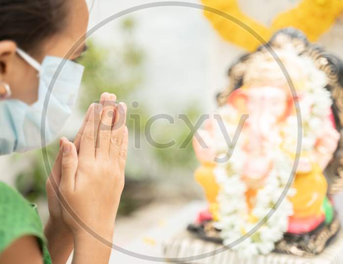 Girl Kid In Medical Mask Praying By Closing Eyes In Front Of Lord Ganesha During Ganesha Or Vinayaka Chaturthi Festival - Concept Of Festival Celebrations During Covid-19 Or Coronavirus Pandemic.