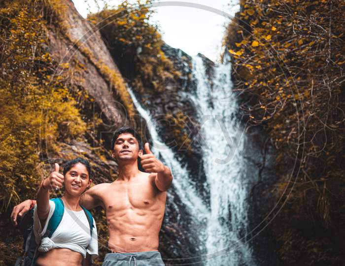 Young Couple Enjoying The Beautiful Waterfall View And Showing Thumbs Up In The Camera.