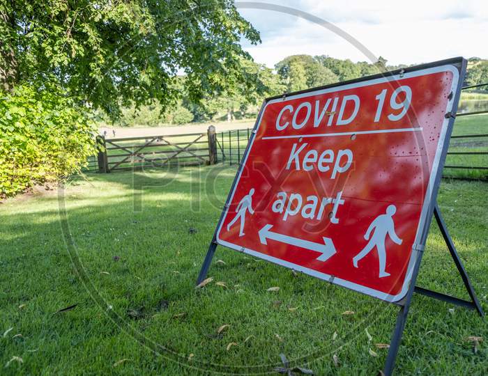 A Red Sign Warning About The Coronavirus Pandemic And Social Distancing In A Field At An Outdoor Event