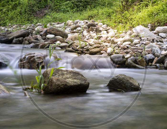 A Rock With Small Plantation Situated In The Middle Of A River Captured In Slow Shutter Speed.