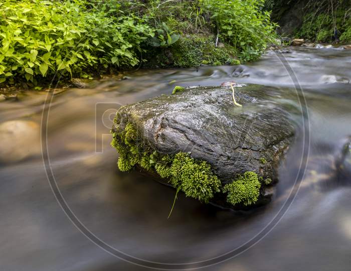 A Rock With Small Plantation Situated In The Middle Of A River Captured In Slow Shutter Speed.