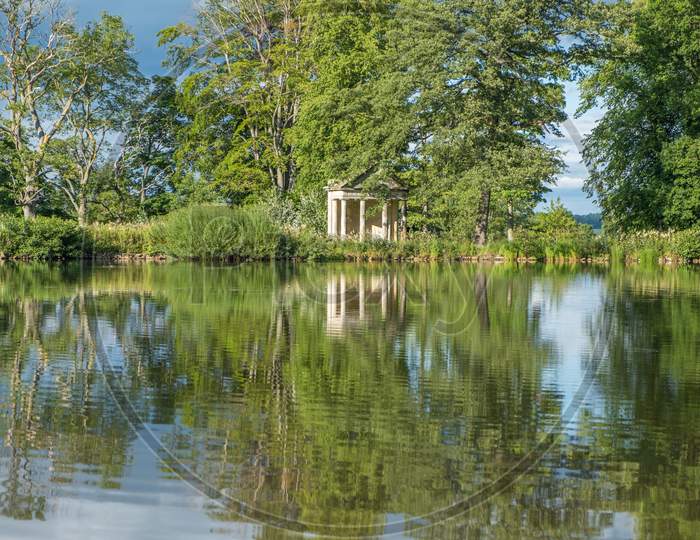A View Across A Beautiful Lake With Reflections Of Trees Towards An Old Abandoned Structure In The Middle Of A Rural Countryside Scene