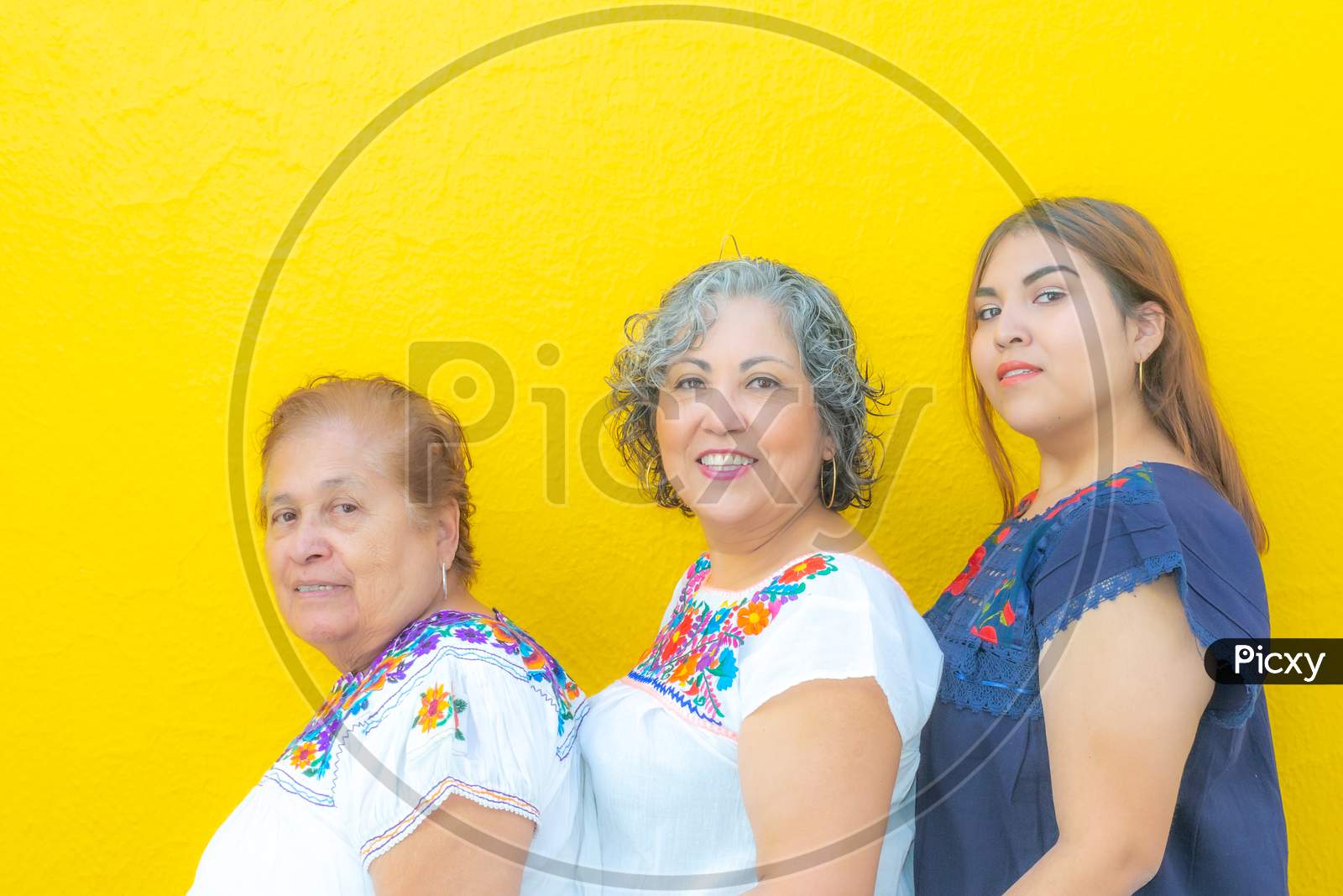 Grandmother, Daughter And Granddaughter With Floral Print Blouses In A Row Looking At The Camera, Three Generations Of Mexican Women On A Yellow Background