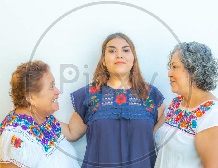 Mother And Daughter Looking At Each Other And  Granddaughter In The Middle Of Them, Three Generations Of Mexican Women Smiling With Floral Print Blouses On A White Background