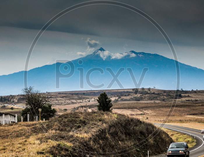 Mexican Landscape With A Curved Asphalt Highway Next To A Small Hill, Cars Driving With The Colima Volcano In The Background, Cloudy Day With Abundant Clouds In The State Of Jalisco, Mexico