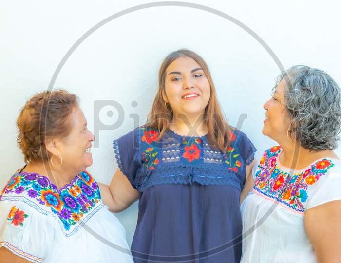 Grandmother And Mother Looking At The Granddaughter, Three Generations Of Mexican Women Smiling With Floral Print Blouses On A White Background
