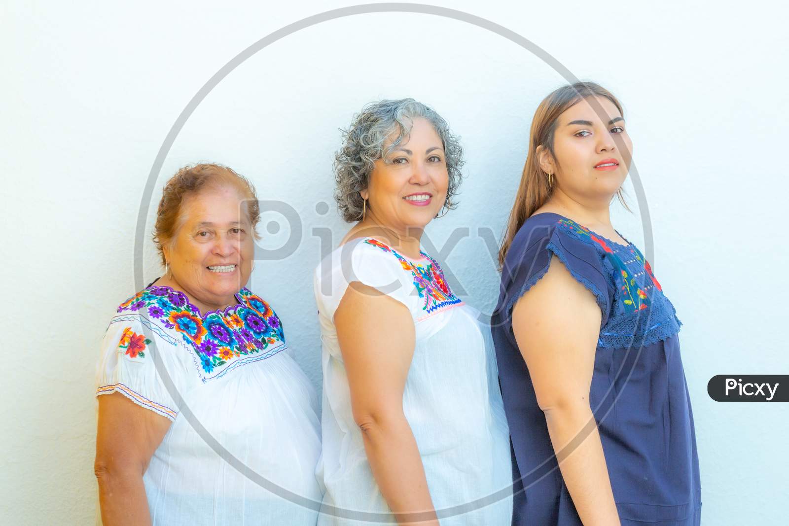Three Generations Of Smiling Mexican Women With Floral Print Blouses In A Row Looking At The Camera On A White Background