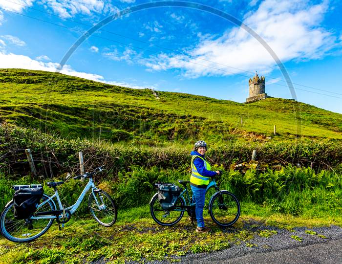 Female Cyclist Having A Break With The Doonagore Castle Tower In The Background In The Coastal Town Of Doolin, Wild Atlantic Way, Wonderful Sunny Day In County Clare In Ireland