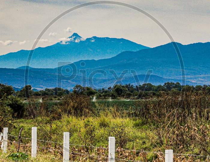 Mexican Landscape With A Field With A Fence With Barbed Wire, Mountains And The Colima Volcano In The Background, Cloudy Day With Abundant Clouds In The State Of Jalisco, Mexico