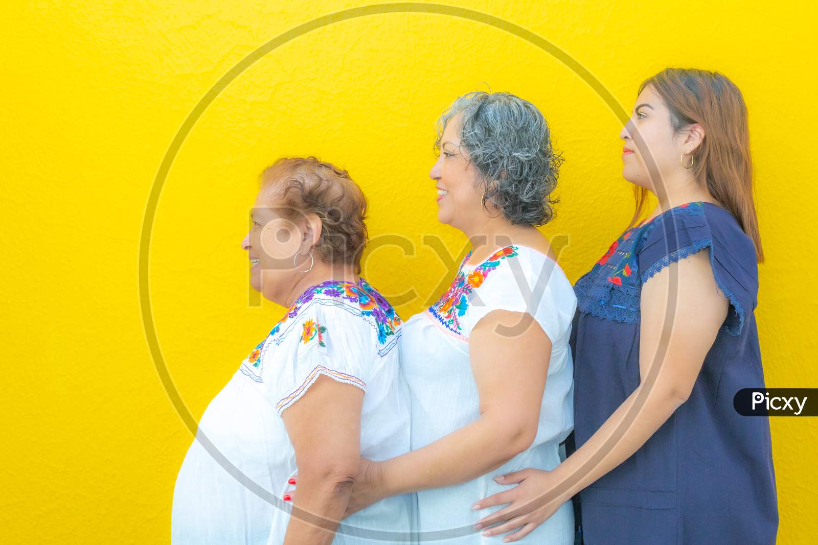 Side View Profiles Of Grandmother, Daughter And Granddaughter, Three Generations Of Mexican Women Smiling With Floral Print Blouses In A Row On A Yellow Background