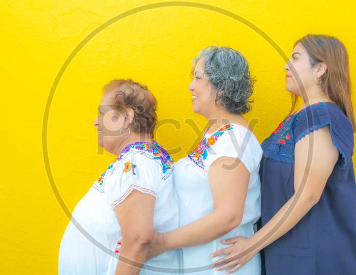 Side View Profiles Of Grandmother, Daughter And Granddaughter, Three Generations Of Mexican Women Smiling With Floral Print Blouses In A Row On A Yellow Background