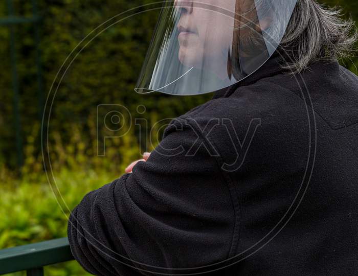 Mature Woman With Glasses In Her Garden Observing The Street With A Medical Protection Screen Or Face Shield To Prevent The Spread Of Covid-19 Amid The Outbreak Of Coronavirus, New Lifestyle