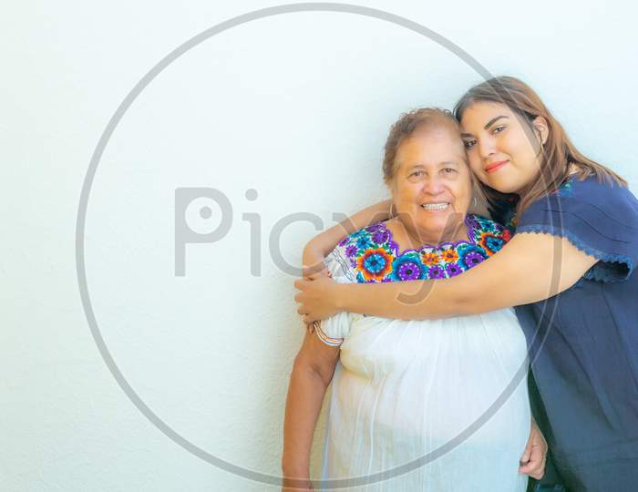 Granddaughter Hugging Her Grandmother, Two Smiling Mexican Women On A White Background, Space For Text