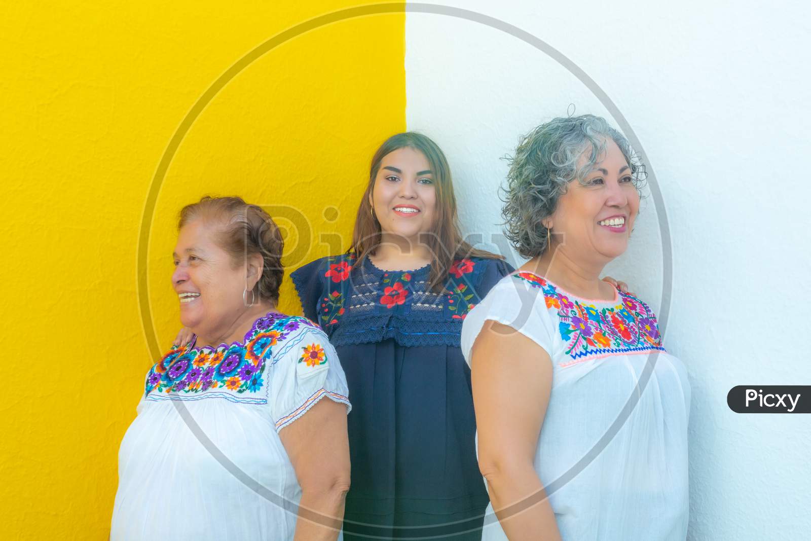 Granddaughter Between Mother And Grandmother Very Cheerful, Three Generations Of Mexican Women Smiling With Floral Print Blouses On A White And Yellow Background
