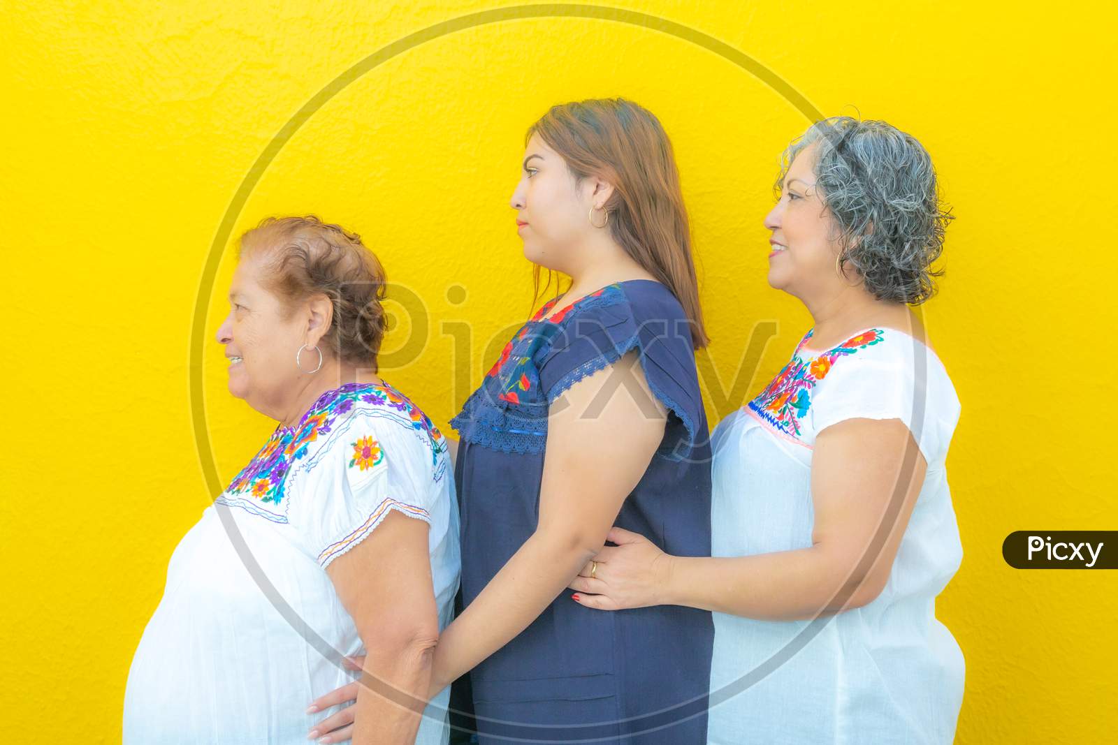 Side View Profiles Of Three Generations Of Mexican Women Smiling With Floral Print Blouses In A Row, Grandmother, Granddaughter And Daughter On A Yellow Background