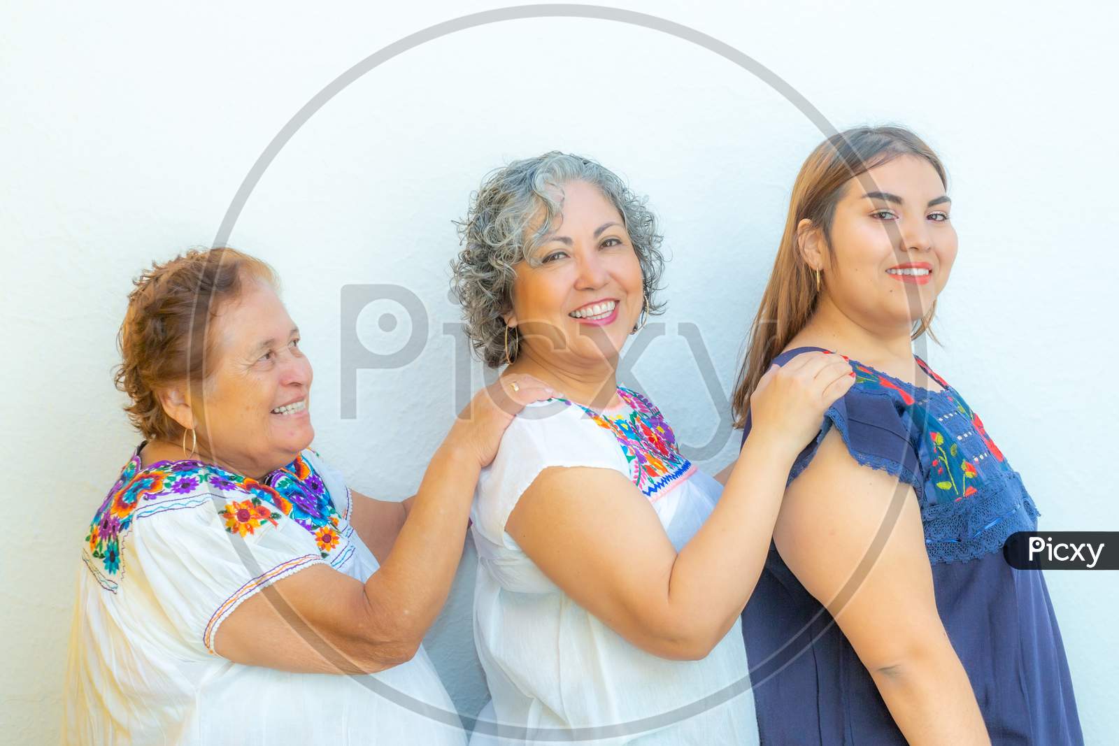 Three Generations Of Smiling Mexican Women With Blouses With Floral Patterns In A Row Holding Their Shoulders Against A White Background