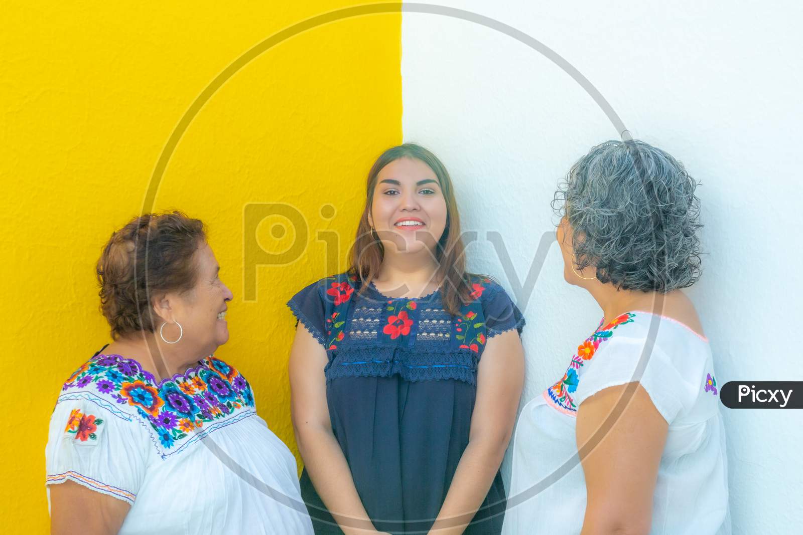 Mother And Grandmother Looking At The Very Cheerful Granddaughter, Three Generations Of Mexican Women Smiling With Floral Print Blouses On A White And Yellow Background