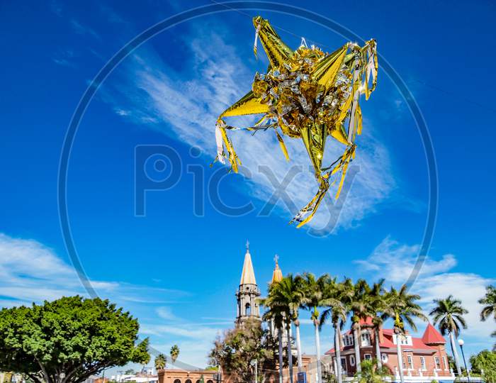 Mexican Peñata Against A Blue Sky, With A Church And Palmtrees In The Background
