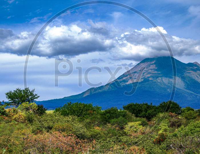 Mexican Landscape With A Hill With Green Vegetation And The Colima Volcano In The Background, Sunny Day With A Blue Sky And Abundant Clouds In The State Of Jalisco Mexico