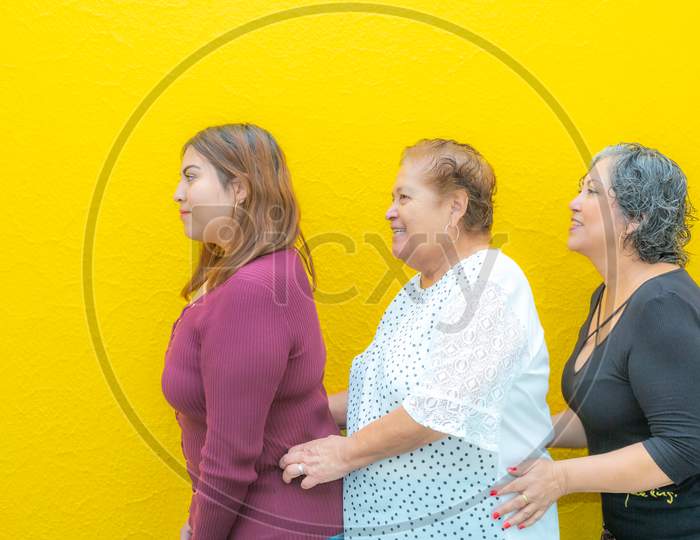 Side View Profiles Of Granddaughter, Grandmother And Daughter Taken From The Waist, Three Generations Of Mexican Women Smiling With Casual Clothes In A Row On A Yellow Background.
