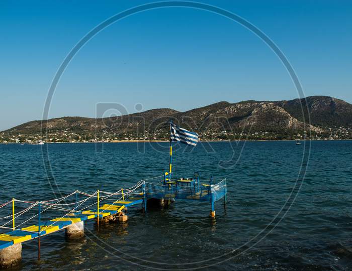 Greek Flag Waves At A Wooden Platform Over The Sea Water Leading To A Table With Three Chairs For Dinner During Summertime Next To The Beach