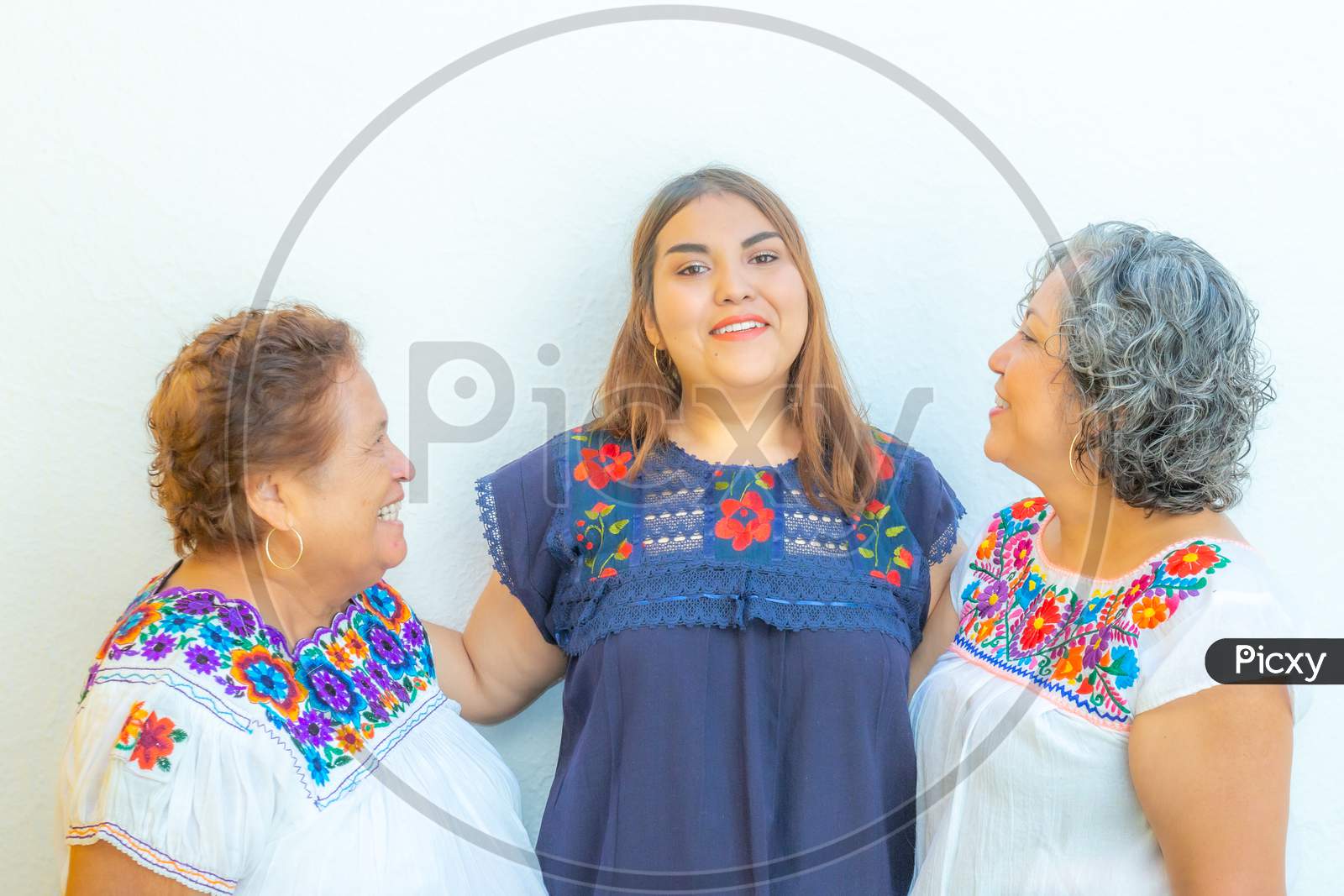 Grandmother And Mother Looking At The Granddaughter, Three Generations Of Mexican Women Smiling With Floral Print Blouses On A White Background