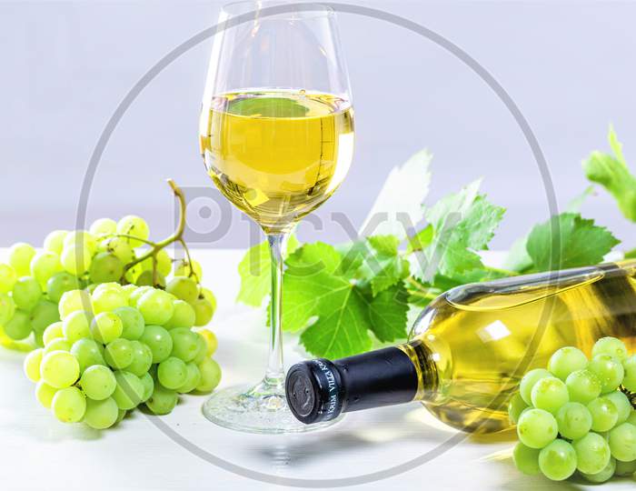 White Wine In A Glass With A Full Bottle, Grapes And Leaves On A White Wooden Background