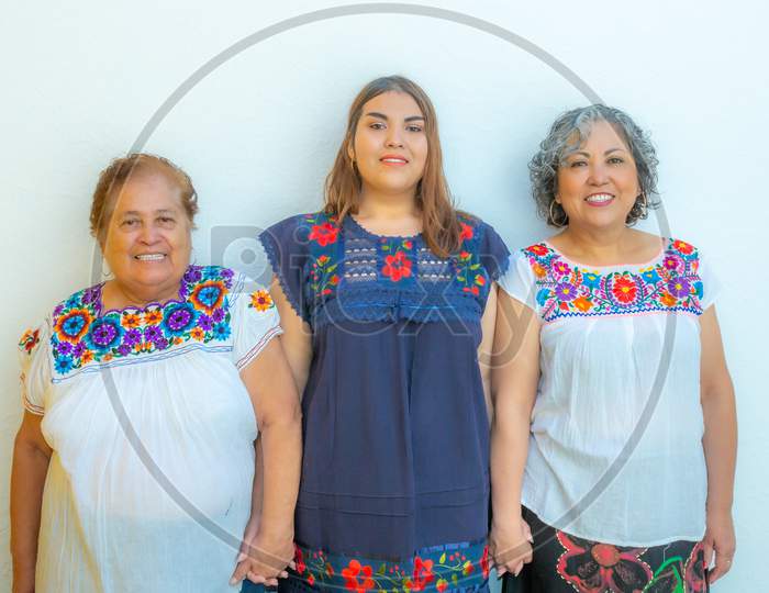 Three Generations Of Latin Mexican Women Smiling, Grandmother, Granddaughter And Daughter With Floral Printed Blouses Holding Hands Looking At The Camera With A White Background