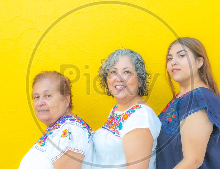 Grandmother, Daughter And Granddaughter With Floral Print Blouses In A Row Looking At The Camera, Three Generations Of Mexican Women On A Yellow Background