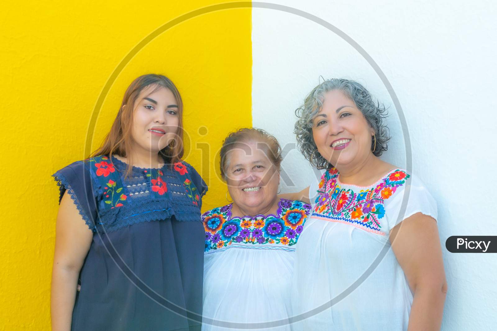 Grandmother Between Daughter And Granddaughter Very Cheerful, Three Generations Of Mexican Women Smiling With Floral Printed Blouses On A White And Yellow Background