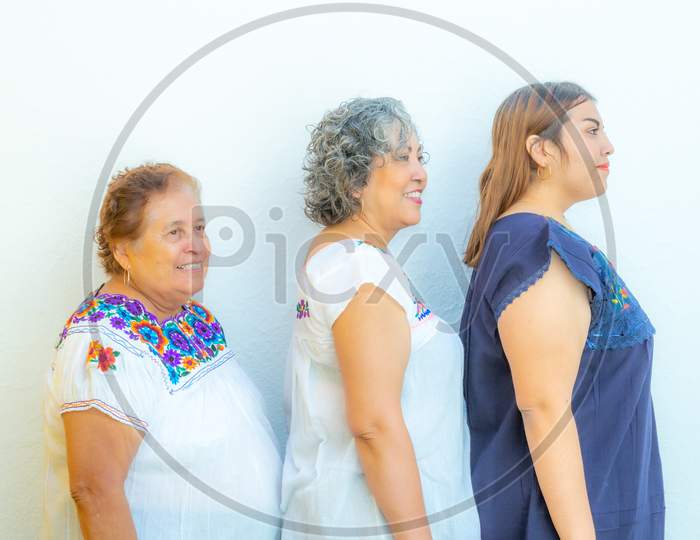 Three Generations Of Smiling Mexican Women With Floral Print Blouses In A Row, Grandmother, Daughter And Granddaughter On A White Background