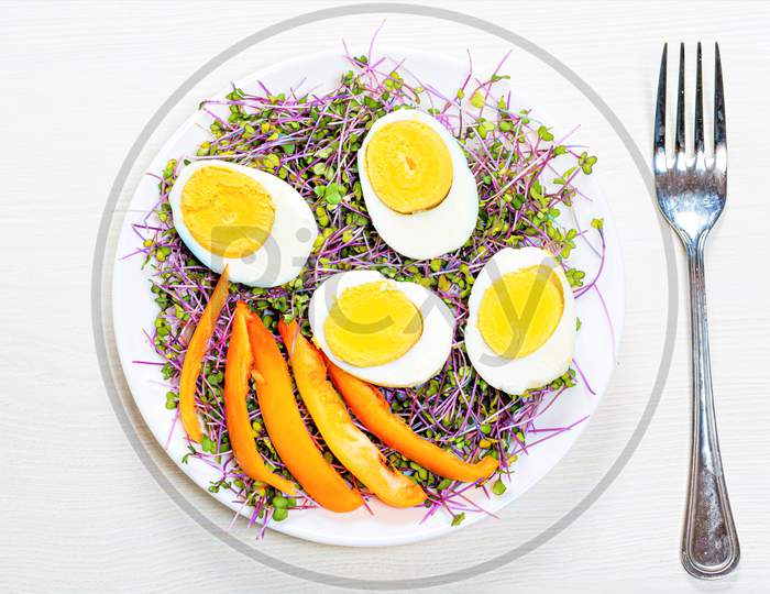 Top View Of Boiled Eggs With Micro Greens And Pieces Of Bell Pepper In A Plate On A White Wooden Background With A Fork